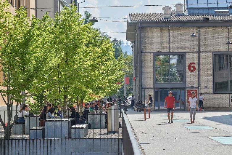 VonRoll – Betontische und -bänke, Studierende sitzen bei Sonnenschein im Schatten der Bäume und diskutieren, oder schlendern über das Areal